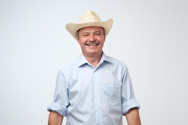 Reifer Mann, der seinen Cowboyhut anpasst und über Witz lacht, während er vor grauem Hintergrund steht