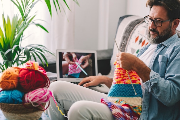 Reifer Mann, der Wolle mit Nadel strickt, während er sich zu Hause ein Online-Tutorial auf dem Laptop ansieht. Mann, der gestrickte Wolle beim Entspannen des Sofas betrachtet. Kaukasischer Mann lernt online stricken