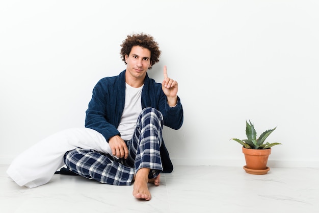 Reifer Mann im Pyjama, der auf Hausboden sitzt und Nummer eins mit Finger zeigt.