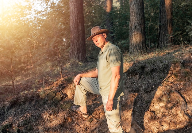 Reifer Mann mit Hut, der im Sommerwald mit Bäumen und Sonnenlicht spazieren geht, der für die Kamera posiert, niedliche männliche Wandern