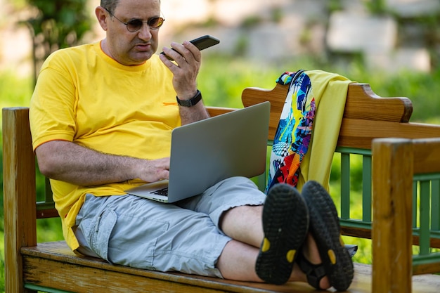 Reifer Mann mit Laptop und Smartphone, der draußen im grünen Home-Office-Konzept des Gartens arbeitet