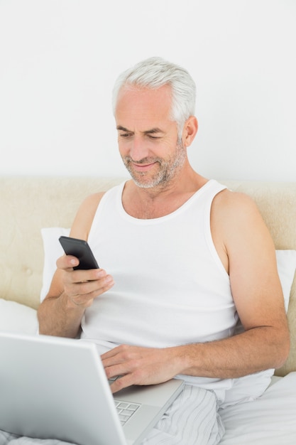 Reifer Mann mit Mobiltelefon und Laptop im Bett