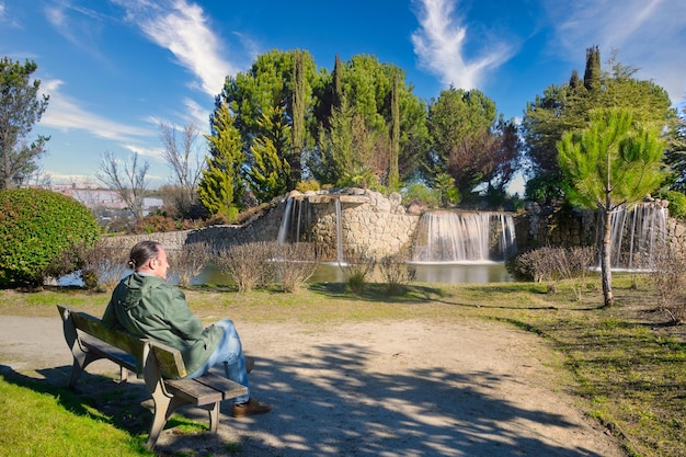 Reifer Mann sitzt auf einer Bank vor einigen Wasserfällen in einem Park in Arroyomolinos, Madrid (Spanien)