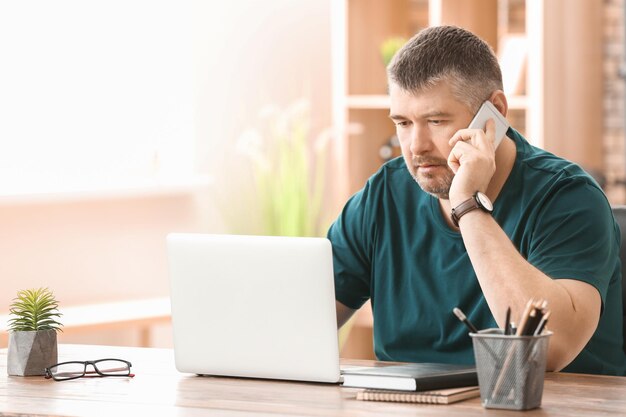 Reifer Mann telefoniert, während er drinnen mit Laptop arbeitet