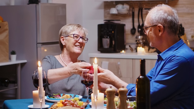 Reifer Mann überraschende Frau mit Geschenk während des Fastenessens, das am Tisch in der Küche sitzt. Fröhliches, fröhliches älteres Ehepaar, das zu Hause zusammen isst, das Essen genießt und ihr Jubiläum feiert?