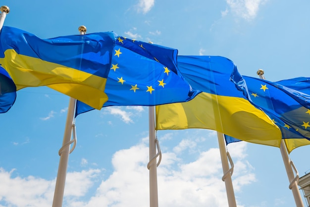 Reihe von Fahnenmasten mit den Fahnen der Europäischen Union und der Ukraine, die vom Wind auf blauem Himmel fladdern