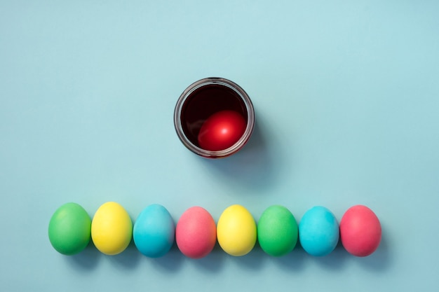 Foto reihe von farbigen ostereiern und einer tasse voll von eifarbe auf pastellblauem hintergrund. flach liegen.