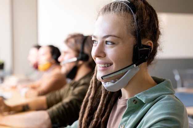 Reihe von Kundenbetreuern mit Headsets, die an Schreibtischen in einem modernen Callcenter sitzen, mit einer jungen Operatorin vor der Tür