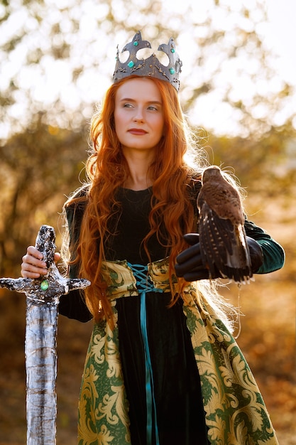 Reina con cabello rojo con un vestido verde con una corona y una espada cerca de un caballo con un pájaro