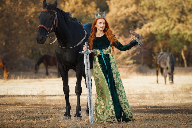 Reina con cabello rojo con un vestido verde con una corona y una espada cerca de un caballo con un pájaro