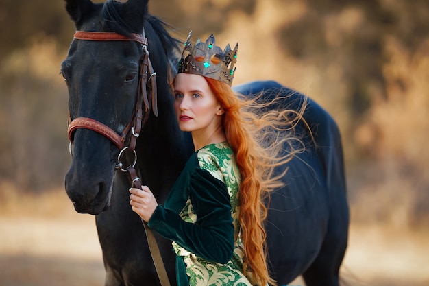 Reina con el pelo rojo con un vestido verde con una corona y cerca de un caballo