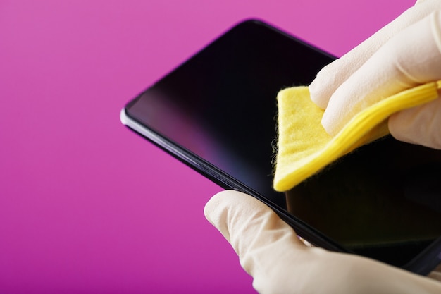 Reinigen eines Smartphones mit einer gelben Serviette von Schmutz und Viren in Gummihandschuhen auf einer rosa Oberfläche