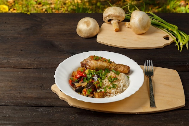 Reis mit Gemüse und Hähnchenschenkel auf einem handgefertigten Holzbrett neben Pilzen und Frühlingszwiebeln auf einem Holztisch