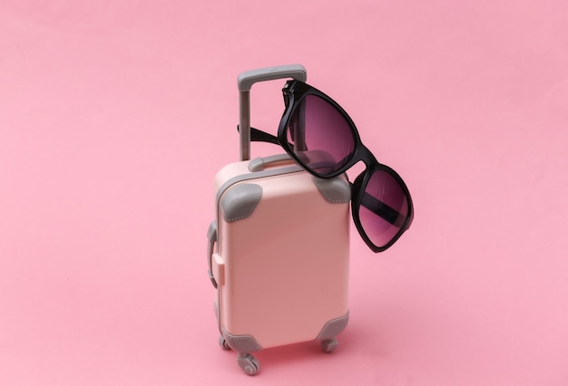 Reise- oder Reisekonzept. Mini Reisekoffer aus Kunststoff mit Sonnenbrille auf rosa Hintergrund. Minimaler Stil