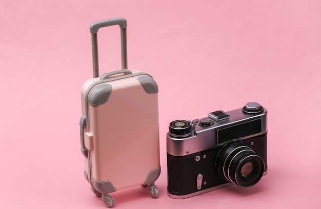 Reise- oder Reisekonzept. Mini-Reisekoffer aus Kunststoff und Kamera auf rosafarbenem Pastellhintergrund. Minimaler Stil