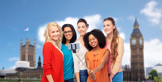 Reise-, Tourismus- und Menschenkonzept - internationale Gruppe glücklich lächelnder verschiedener Frauen, die mit Smartphone auf Selfie-Stick vor dem Hintergrund des Londoner Big Ben Tower fotografieren