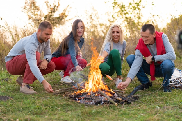 Reise-, Tourismus-, Wander-, Picknick- und Personenkonzept - Gruppe glücklicher Freunde, die Würste am Lagerfeuer braten