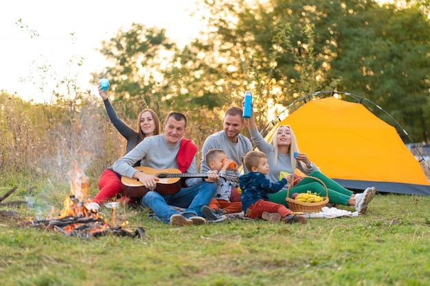 Reise-, Tourismus-, Wander-, Picknick- und Personenkonzept - Gruppe glücklicher Freunde mit Zelt und Getränken, die auf dem Campingplatz Gitarre spielen