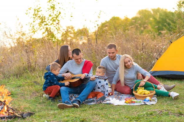 Reise-, Tourismus-, Wanderungs-, Picknick- und Leutekonzept - Gruppe glückliche Freunde mit dem Zelt und Getränken, die Gitarre am Kampieren spielen