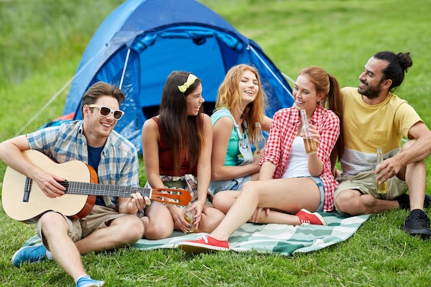 Reise-, Tourismus-, Wanderungs-, Picknick- und Leutekonzept - Gruppe glückliche Freunde mit dem Zelt und Getränken, die Gitarre am Kampieren spielen