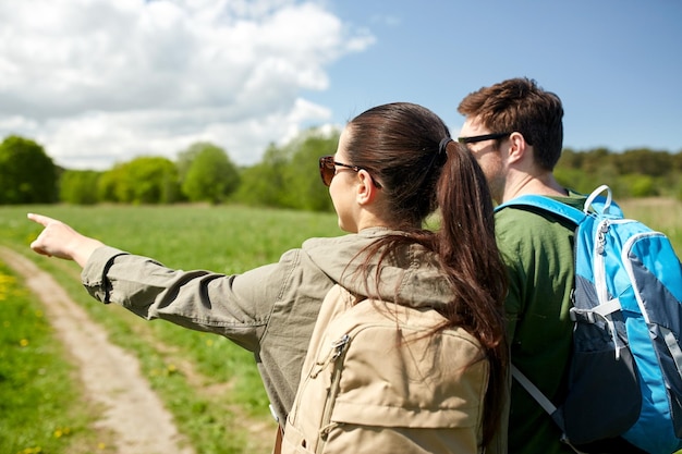 Reise-, Wander-, Rucksackreisen-, Tourismus- und Personenkonzept - glückliches Paar mit Rucksäcken, das auf der Landstraße spaziert und mit dem Finger auf etwas zeigt