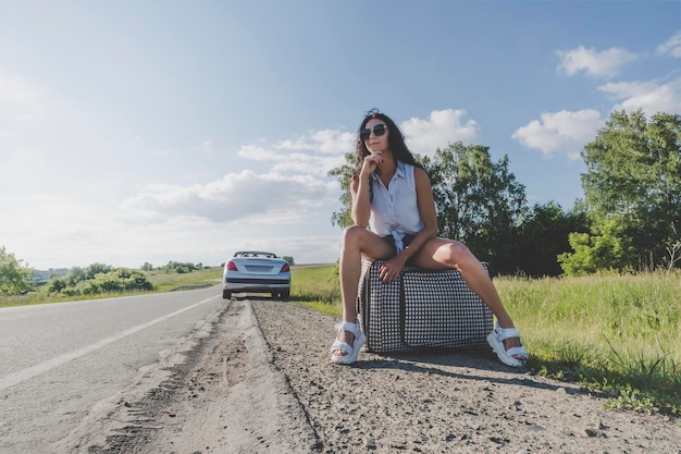 Reiseabenteuer Teenager-Reisekonzept Frau in Denim-Shorts Top-Koffer mit Koffer auf Rädern per Anhalter sitzend und entspannend während der Reise