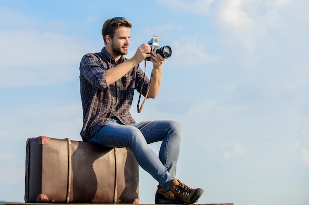 Reiseblog Mann sitzt auf Koffer Gutaussehender Typ Reisender Retro-Kamera Kerl im Freien mit Vintage-Koffer Fotojournalist-Konzept Reisen mit Gepäck Reiseblogger Vlog-Aufnahmen Urlaubszeit