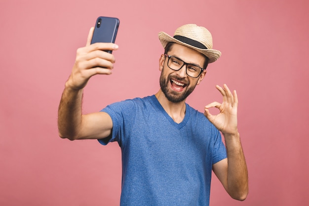 Reisekonzept. Porträt des fröhlichen jungen Mannes im Strohhut, der selfie mit Smartphone lokalisiert über rosa Hintergrund nimmt.