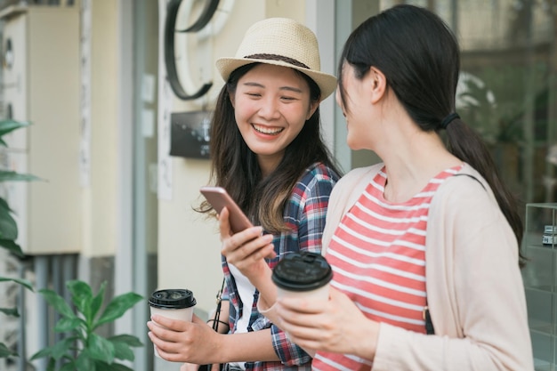 Reiselebensstil an sonnigen Sommertagen. Freundinnen stehen draußen mit Smartphone und Einweg-Pappbecher in den Händen. Zwei Mädchen lachen und teilen sich ihr Handy, während sie am Fenster Kaffee trinken