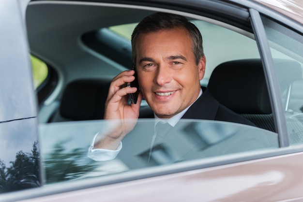 Reisen mit Komfort. Glücklicher reifer Geschäftsmann, der auf dem Handy spricht und lächelt, während er auf dem Rücksitz eines Autos sitzt