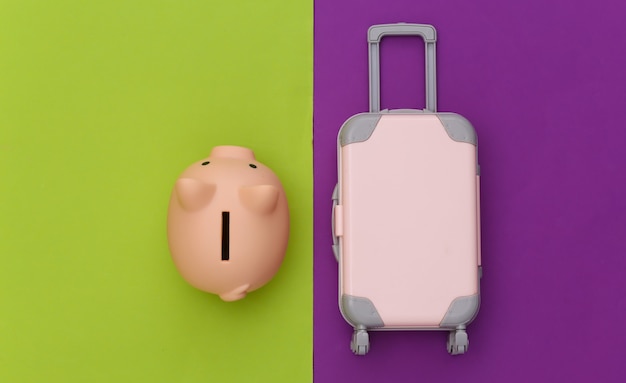 Reisen Sie flach. Mini Reisekoffer aus Kunststoff, Sparschwein auf grünem lila Hintergrund. Minimaler Stil. Ansicht von oben
