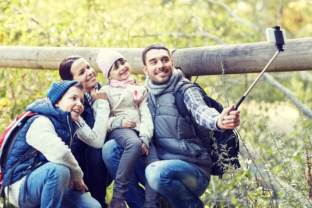 Reisen, Tourismus, Wandern, Technologie und Menschen Konzept - glückliche Familie mit Rucksäcken fotografieren mit Smartphone und Selfie-Stick im Wald