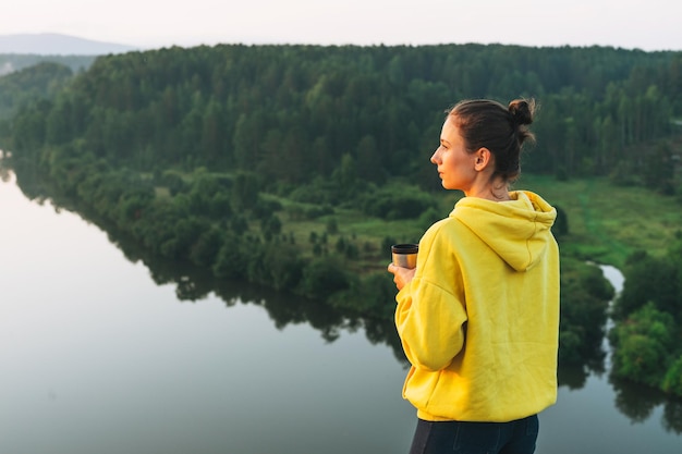 Reisende der jungen Frau im gelben Hoodie mit Tasse Kaffee am Morgen bei Sonnenaufgang auf schöner Aussicht