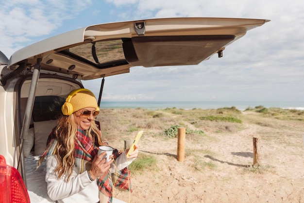Reisende Frau, die sich an einem ruhigen Küstenort mit Handy an einen Van lehnt, hören Sie Musik mit Kopfhörern Reisecamper digitale Nomaden