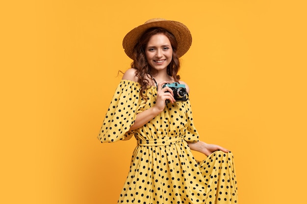 Reisende Frau in Strohhut und Sommerkleid posiert mit Retro-Kamera auf Gelb