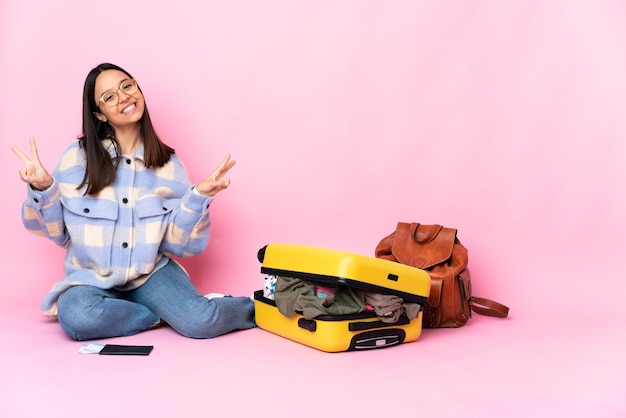 Reisende Frau mit einem Koffer, der auf dem Boden sitzt und Siegeszeichen mit beiden Händen zeigt