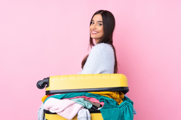 Reisende Frau mit einem Koffer voller Kleidung über isolierte rosa Wand lachen