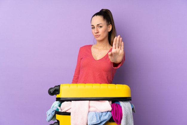Reisende Frau mit einem Koffer voller Kleidung über isolierter lila Wand, die Stoppgeste mit ihrer Hand macht