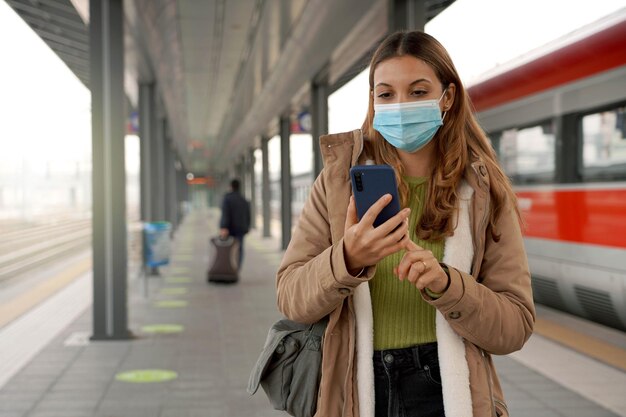 Reisende Frau mit Mundschutz am Bahnhof Ticket online kaufen mit ihrem Handy im Freien