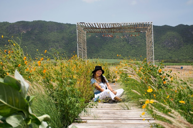 Reisende thailändische Frauen sitzen und posieren Porträt auf einer Holzbrücke im Garten im Freien und reisen, besuchen Sie, entspannen Sie sich in einer Blumenwiese mit Wind- und Berghintergrund in Kanchanaburi Thailand