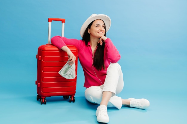 Reisende Touristin in Freizeitkleidung und weißem Hut, die Geld in der Hand hält und am roten Koffer sitzt, isoliert auf blauem Hintergrund. Passagierin, die ins Ausland reist, um einen Wochenendausflug zu machen