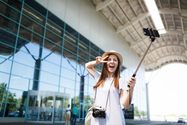 Reisende Touristin mit Retro-Vintage-Fotokamera zeigt Victory-Zeichen, macht Selfie auf dem Handy mit egoistischem Einbeinstativ am Flughafen