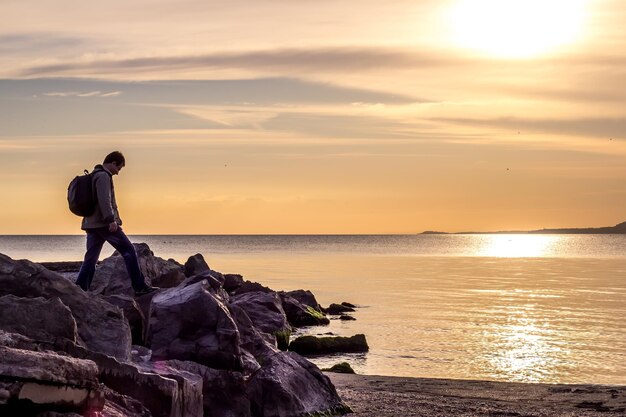 Reisender, der auf Felsen gegen Meer, Sonnenaufgang oder Sonnenuntergang friedliche Landschaft geht