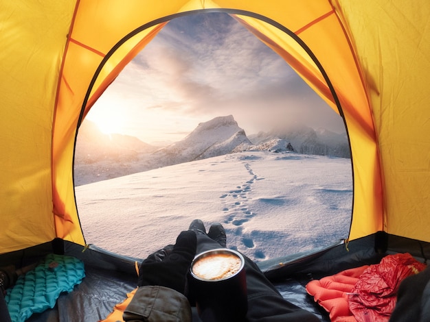 Reisender, der eine Kaffeetasse hält und den Blick auf den Sonnenaufgang auf einem schneebedeckten Berg in einem gelben Zelt genießt