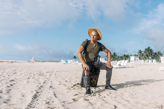 Reisender gutaussehender Hipster-Mann, der am schönen Strand mit Gepäck sitzt und eine atemberaubende Aussicht genießt. Trägt einen Hut und eine zerrissene Jeans