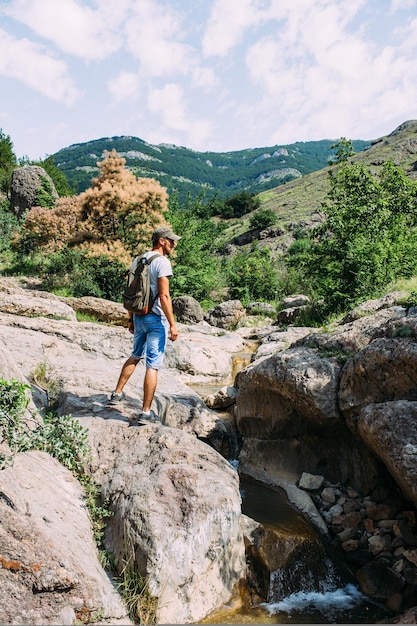 Reisender junger Mann, der allein an einem Bergfluss mit felsiger Sommerlandschaft spazieren geht?