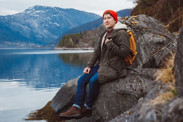 Reisender Mann mit gelbem Rucksack sitzt am Ufer vor dem Hintergrund von Berg und See