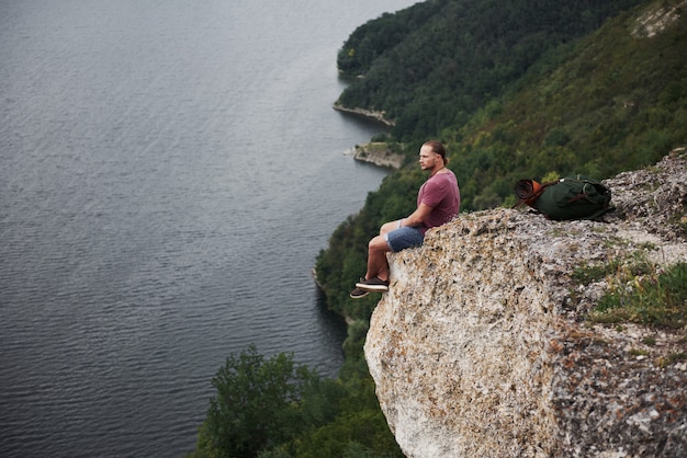 Reisender mit Rucksack, der oben auf Berg sitzt und Blick über der Wasseroberfläche genießt. Freiheit und aktiver Lebensstil Konzept