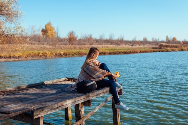 Reisender mit Rucksack, der sich am Herbstfluss bei Sonnenuntergang mit Blättern entspannt. Junge Frau, die auf Pier sitzt und Landschaft und Wetter bewundert. Aktiver Lebensstil