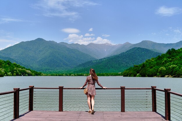 Reisendes Mädchen, das allein am Rand des Piers steht und auf See und Berge starrt. Genießen Sie das schöne Freiheitsmoment und die ruhige, friedliche Atmosphäre in der Natur. Rückansicht